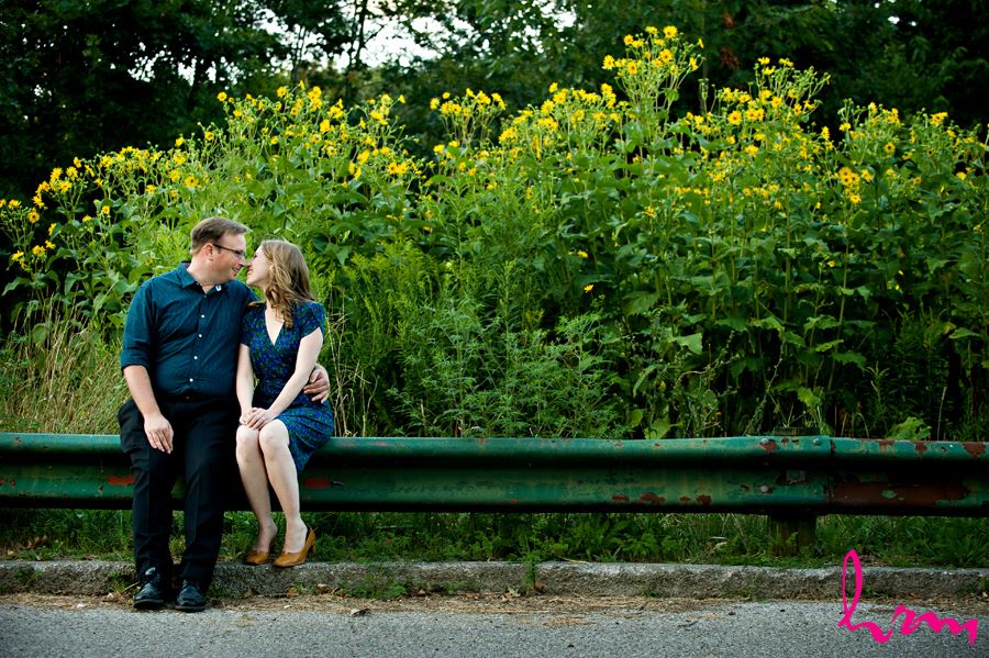 Beautiful engaged couple sitting on railing with yellow flowers behind them