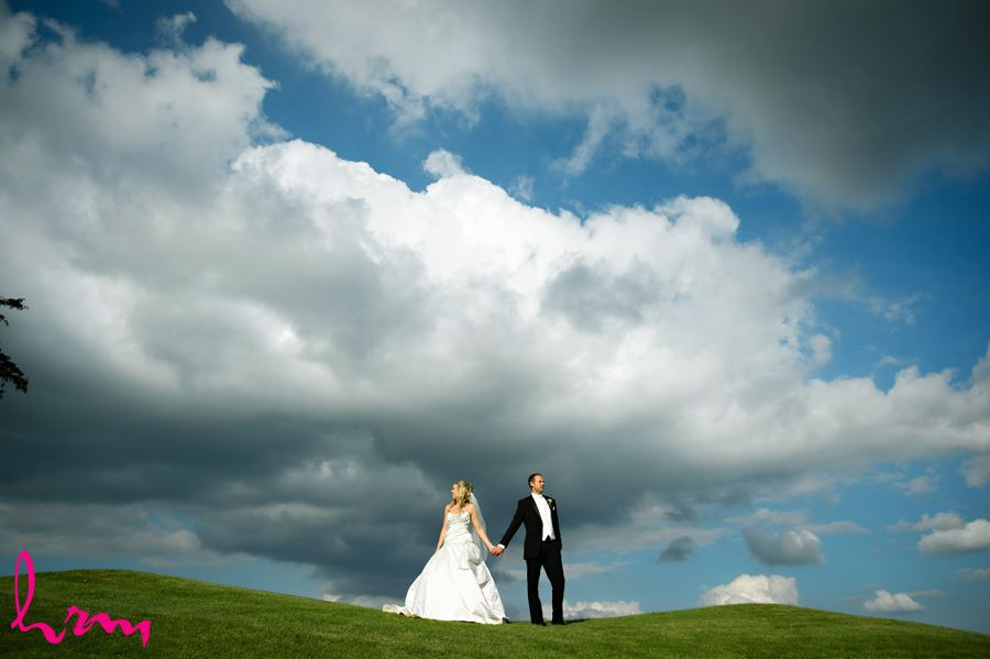 Bride and groom pose on a green hill with bright blue sky andbig white clouds
