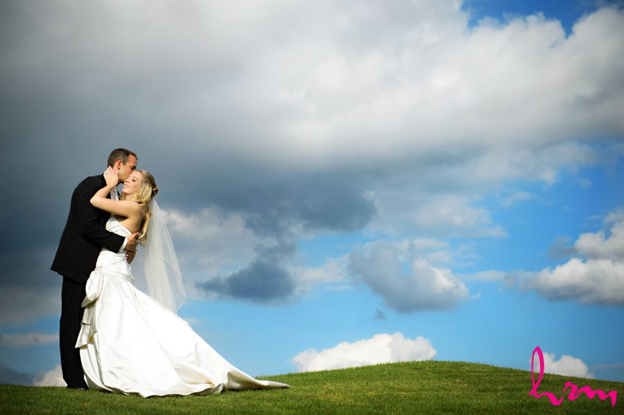 Groom kissing brides forehead on a green hill with bright blue background and big white clouds in London Ontario